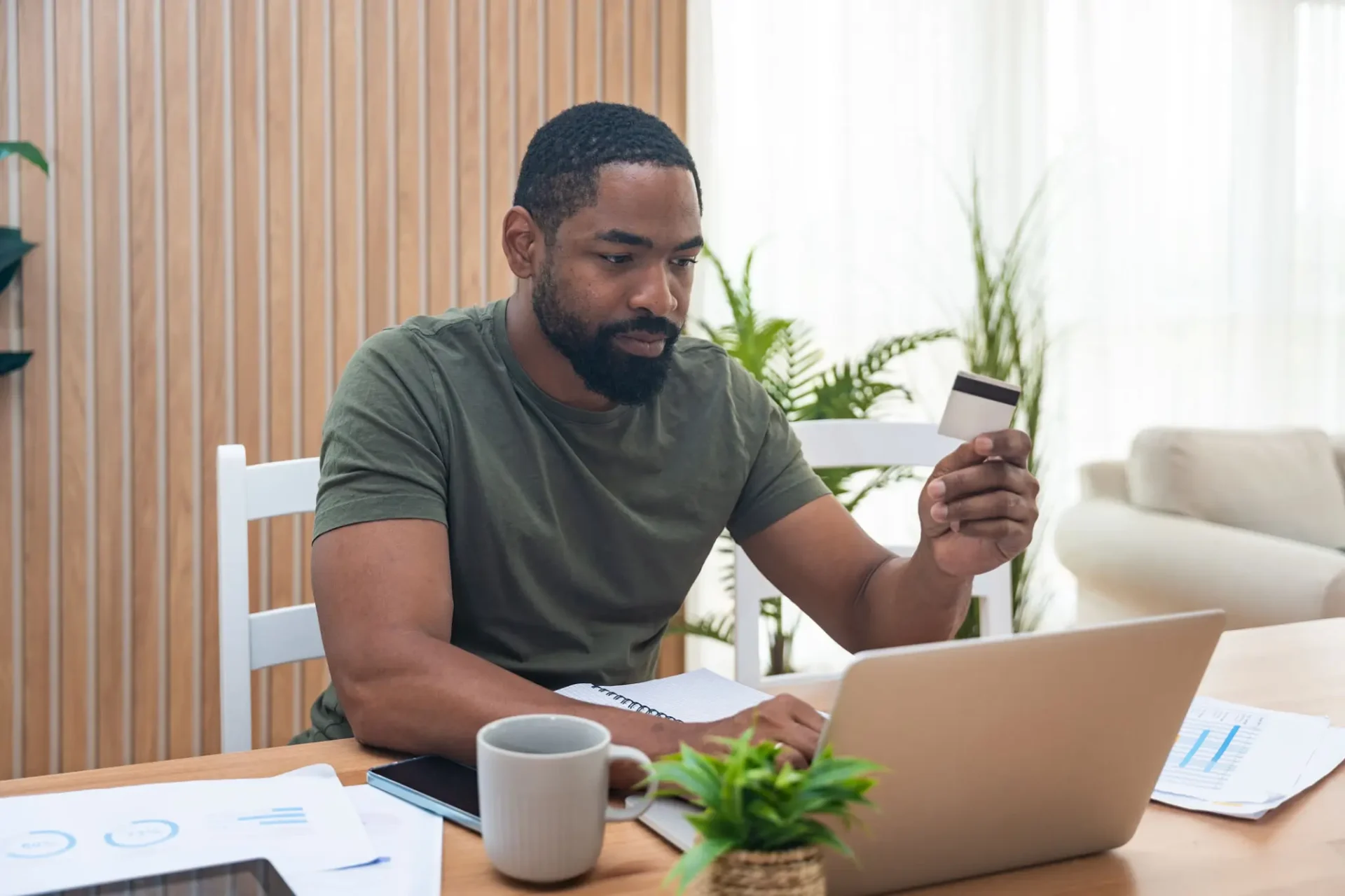 Man working on laptop while holding a smartphone, surrounded by plants and a coffee cup.
